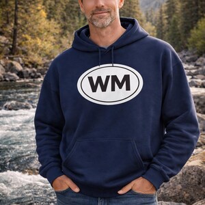 May include: A navy blue hooded sweatshirt featuring a white oval design with the black letters "WM". The sweatshirt has a front pocket and a drawstring hood. The person is wearing blue jeans. The background is a river.