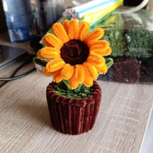 Peut inclure: Un tournesol décoratif fait à la main dans un petit pot. Le tournesol a des pétales orange vif et un centre brun foncé. Le pot est d'un brun rougeâtre profond et a une texture côtelée. La fleur est faite d'un matériau doux et duveteux.