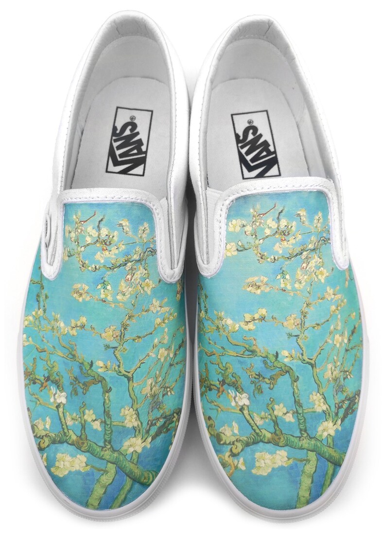 Puede incluir: Un par de zapatos de lona blancos sin cordones con un estampado de una pintura de una rama de almendro en flor contra un cielo azul. Los zapatos tienen la palabra "VANS" impresa en el interior de cada zapato.