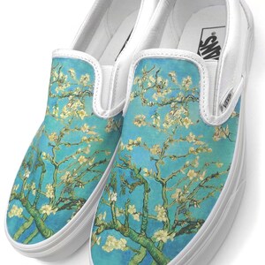Puede incluir: Un par de zapatos de lona blancos sin cordones con un estampado floral azul y blanco. El estampado presenta una pintura de un almendro en flor de Vincent van Gogh.