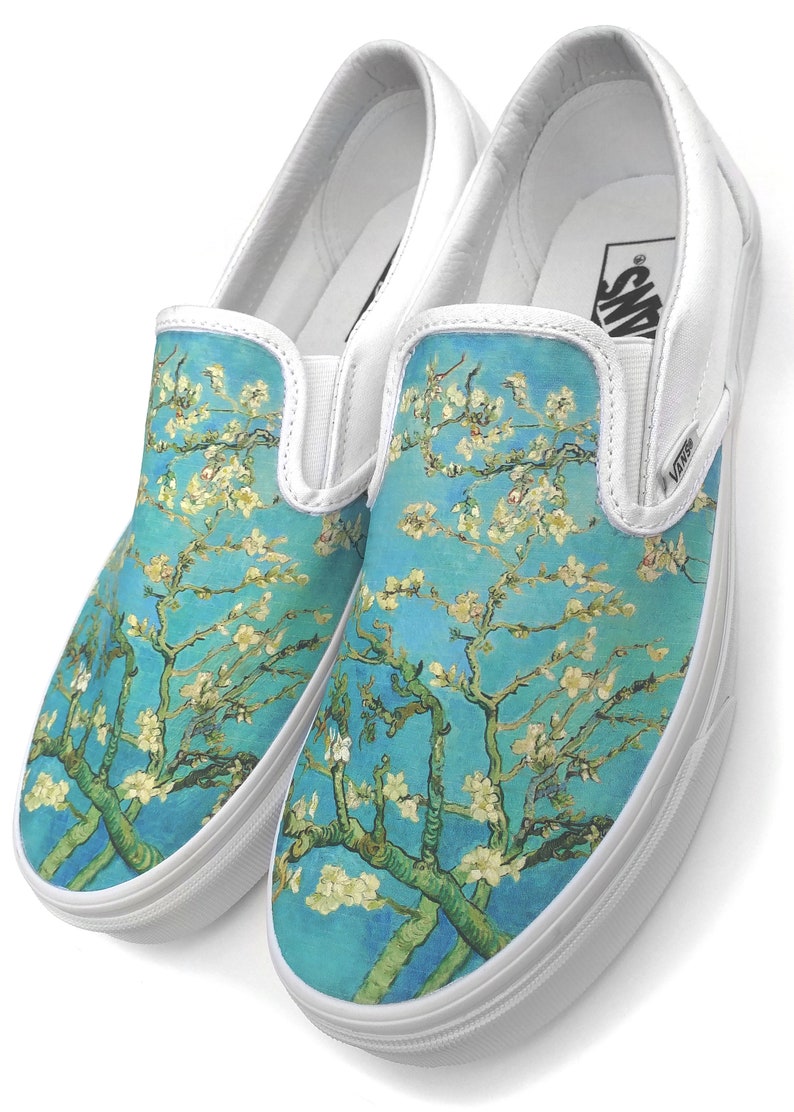 Puede incluir: Un par de zapatos de lona blancos sin cordones con un estampado floral azul y blanco. El estampado presenta una pintura de un almendro en flor de Vincent van Gogh.