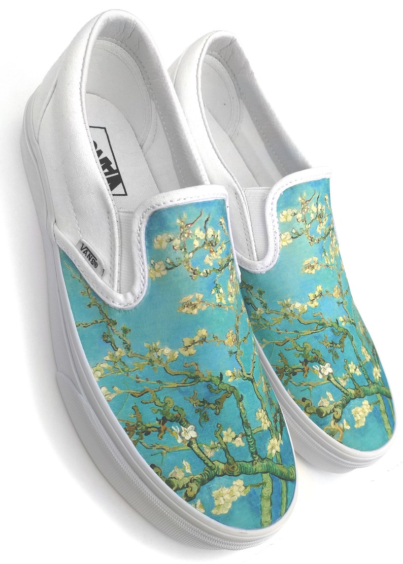 Puede incluir: Zapatillas Vans blancas de deslizamiento con un estampado floral azul y blanco. El estampado presenta una representaci&oacute;n estilizada de una rama de almendro en flor, que recuerda a la obra de arte de Vincent van Gogh.