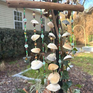 May include: A handcrafted seashell wind chime. The chime features a natural wooden beam with multiple strands of seashells, wooden beads, and green and clear beads. The seashells are various sizes and colors, creating a coastal aesthetic.