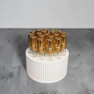 May include: A white cylindrical organizer with vertical ribbed detailing holds nineteen small, clear glass spray bottles. Each bottle is topped with a metallic gold cap and features a clear pump mechanism, arranged in a compact circular formation.