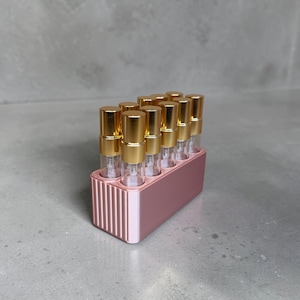 May include: A collection of small perfume or essential oil vials with gold-colored caps, arranged in a pink rectangular holder. The holder has vertical lines on the front. The vials are clear, allowing the contents to be visible.