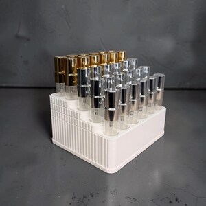 May include: A white tiered display stand holding multiple small, clear glass spray bottles. The bottles feature metallic caps in gold and silver finishes, arranged in rows on the stepped structure of the rectangular plastic organizer.