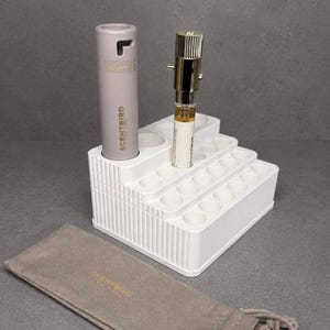 May include: A white tiered plastic organizer with multiple circular slots for small cylindrical items. A light gray Scentbird perfume case and a small glass vial with a gold cap are placed in the slots. A gray fabric pouch with Scentbird text is included.