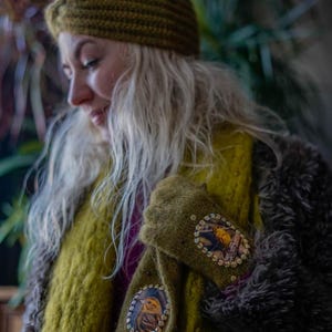 May include: Olive green knitted headband, scarf, and mittens. The mittens feature a circular design with a bird illustration and sequin embellishments. The scarf and headband are a similar shade of green.