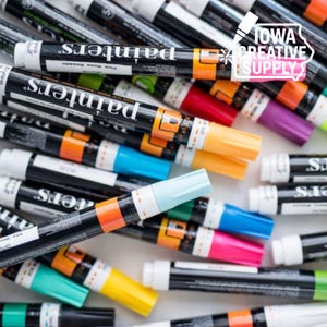 May include: A collection of colorful paint pens with black barrels and white caps. The pens are arranged in a scattered pattern, showcasing various colors like orange, yellow, blue, pink, and purple. The text "painters" is visible on the pens.