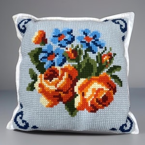 Peut inclure: Un coussin carré avec une housse en tissu bleu clair brodé au point de croix, orné d'un bouquet central de roses orange et de fleurs bleues. Le coussin est bordé d'un liseré blanc et présente des motifs en volutes bleu foncé aux coins.