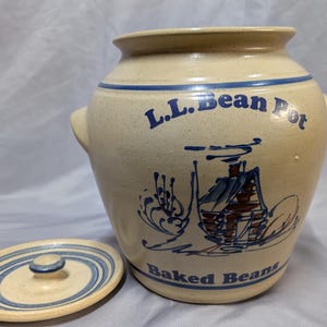 May include: Tan ceramic bean pot with blue accents and the text "L.L. Bean Pot Baked Beans." The pot has a lid with blue stripes and a small handle. The pot features a cabin illustration.