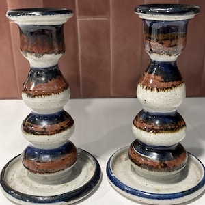 May include: Two ceramic candle holders with a unique design. The holders feature a white base with brown, blue, and black accents. Each has a round base and a top designed to hold a candle. The candle holders are a decorative item.