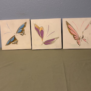 Könnte beinhalten: Set aus drei quadratischen Wandkunstwerken, jedes mit einem stilisierten Schmetterlingsdesign. Die Schmetterlinge haben goldene, blaue, rosa und lila Flügel auf cremefarbenem Hintergrund. Ein dekoratives Wandbild.