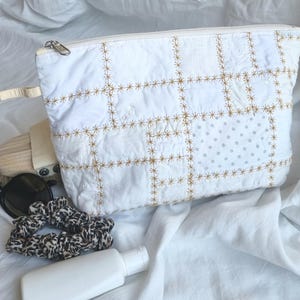 Puede incluir: Un estuche blanco con cremallera que presenta un diseño de patchwork de varios tejidos blancos unidos por bordados dorados en forma de estrella. El estuche va acompañado de gafas de sol negras, un coletero con estampado de leopardo y un frasco blanco.