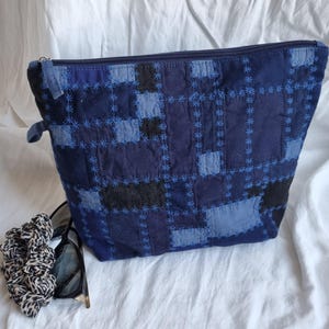 May include: A zippered pouch featuring a patchwork design of various blue and black fabric squares, accented with light blue star-shaped embroidery stitching. Beside the pouch are black-framed sunglasses and a leopard print fabric hair scrunchie.