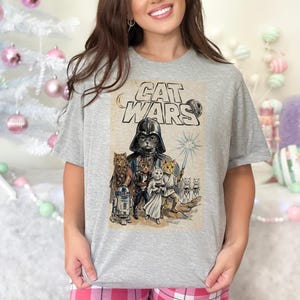Może przedstawiać: Szary melanżowy t-shirt z krótkim rękawem z nadrukiem graficznym i napisem CAT WARS. Wzór przedstawia różne koty przebrane za postacie science-fiction, w tym kota w ciemnym hełmie oraz kota trzymającego świecący miecz świetlny.