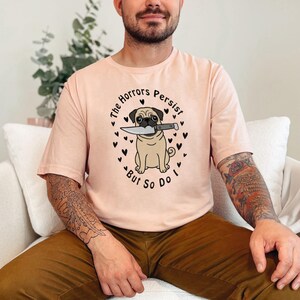 May include: A light peach-colored t-shirt featuring a graphic of a tan pug holding a silver knife in its mouth. The text "The Horrors Persist, But So Do I" is printed in a circular arc around the pug, surrounded by small black heart icons.