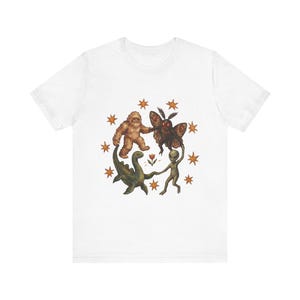 Può includere: Una t-shirt bianca a maniche corte con una grafica centrale che raffigura un Bigfoot marrone, una creatura scura simile a una falena, un dinosauro acquatico verde e un alieno grigio che si tengono per mano in cerchio attorno a un fiore, circondati da stelle arancioni.