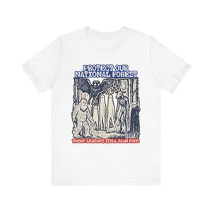 May include: A white t-shirt featuring a dark blue graphic print of a forest scene with a sasquatch, ghosts, a deer-headed figure, and a bat. The text reads PROTECT OUR NATIONAL FOREST at the top and WHERE LEGENDS STILL ROAM FREE in red at the bottom.