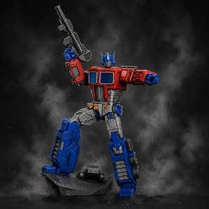 Op de afbeelding: Een rood, blauw en grijs Optimus Prime actiefiguur staand op een donkere rotsbasis. Het figuur houdt een zwart geweer in de opgeheven rechterhand en heeft een zilverkleurig Autobot-insigne op de linkerschouder. Eén voet rust op de rots.