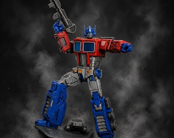 May include: A red, blue, and gray Optimus Prime action figure standing on a dark rock base. The figure holds a black rifle in its raised right hand and features a silver Autobot insignia on its left shoulder. One foot rests on the rock.