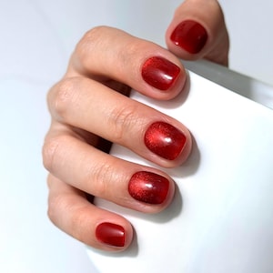 May include: Fingernails painted with a glossy, deep red polish featuring a subtle shimmering effect. The nails are shaped with rounded edges and a smooth, reflective finish, showcasing a vibrant crimson color that catches the light.