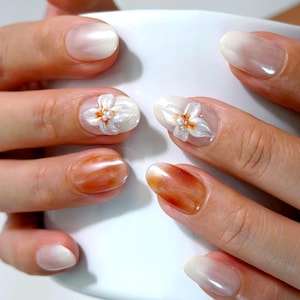 May include: Manicured fingernails featuring a mix of designs including a soft white gradient, a solid warm amber marble effect, and a three-dimensional white floral accent with small pearl-like centers on the middle fingernails.
