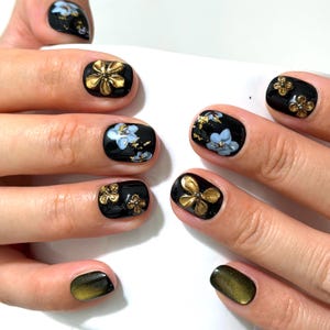 May include: Manicured fingernails featuring a glossy black base coat. The nails are decorated with three-dimensional gold flower charms, hand-painted light blue floral accents, gold leaf flakes, and two nails with a shimmering olive green cat-eye finish.