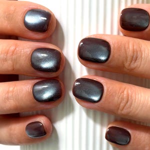 May include: Short, rounded fingernails featuring a dark, metallic charcoal gray polish with a magnetic cat-eye effect. The finish is glossy and reflective, showing a subtle shimmer that shifts in light across the surface of each nail.