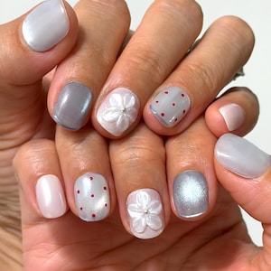 May include: Manicured fingernails featuring a mix of designs including solid light gray, solid pale pink, gray with small red polka dots, and pale pink with a raised white five-petaled flower accent featuring a small silver center bead.