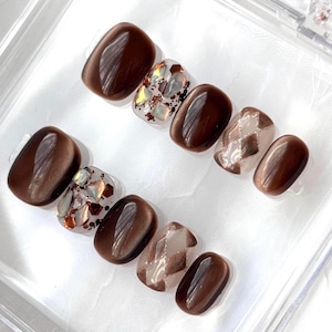 May include: A set of ten press-on nails featuring a deep brown cat-eye finish, clear nails with iridescent chunky glitter flakes, and clear nails with a brown argyle pattern. The nails have a smooth, glossy surface and a rounded square shape.