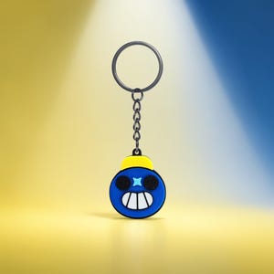 May include: A blue and yellow cartoon bomb keychain with a black chain and keyring. The bomb has a smiling face with white teeth and a star-shaped eye. The background is a gradient of yellow and blue.