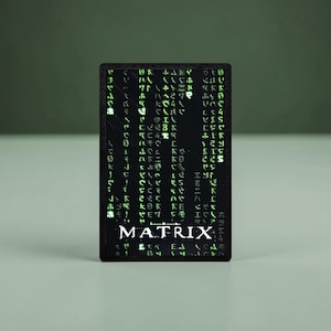 Könnte beinhalten: Eine rechteckige schwarze Karte mit einem vertikalen Muster aus leuchtend grünen digitalen Zeichen und Symbolen. Das Wort MATRIX ist in weißen Großbuchstaben unten in der Mitte der Karte aufgedruckt, mit einer stilisierten horizontalen Linie über dem T.