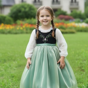 Peut inclure: Une jeune fille porte une robe verte et noire avec un haut blanc à manches longues. La robe présente un corsage noir avec des broderies florales et une jupe vert menthe. La fille sourit et a ses cheveux en deux tresses.