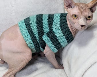 May include: A hairless cat wearing a green and turquoise striped crocheted sweater. The cat is sitting on a white fluffy surface.