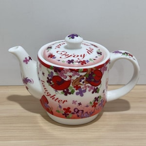 Puede incluir: Tetera de cerámica blanca con tapa, decorada con sombreros rojos, flores moradas y las palabras "Enjoy the laughter". La tetera tiene un pico y un asa curvados.