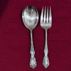 Puede incluir: Una cuchara de servir y un tenedor de servir de cuatro dientes en metal plateado. Ambos utensilios presentan un cuenco y dientes pulidos y reflectantes, con detalles florales y de volutas en relieve a lo largo de los mangos.