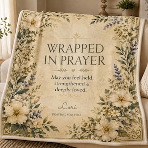 May include: A cream-colored throw blanket featuring a floral border with white and blue flowers. The text reads: WRAPPED IN PRAYER, May you feel held, strengthened & deeply loved., Lori, PRAYING FOR YOU. The design includes decorative flourishes.