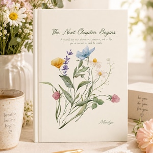 May include: A white hardcover journal featuring a watercolor illustration of a wildflower bouquet. The text reads The Next Chapter Begins, A journal for new adventures, dreams, and a life you've worked so hard to create, and the name Marilyn.