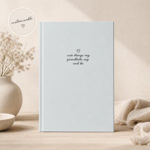 Op de afbeelding: Een lichtblauw notitieboek met de tekst "cute things my grandkids say and do" in zwart schrift, met een hartsymbool erboven. Een kleine, ronde "customizable" sticker staat in de linkerbovenhoek. Het notitieboek ligt op hout.