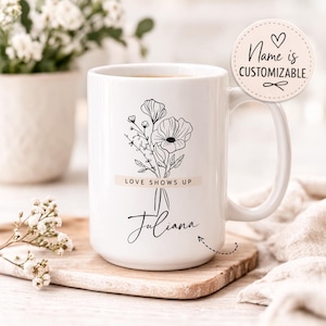Peut inclure: Mug en céramique blanche avec un motif floral noir, le texte "LOVE SHOWS UP" et "Juliana". Un autocollant rond sur la droite indique "Name is Customizable". Le mug est posé sur un dessous de verre en bois.