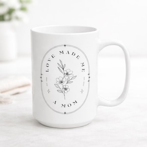 Könnte beinhalten: Weißer Keramikbecher mit großem Henkel. Der Becher hat ein schwarzes Blumendesign in einem Oval mit den Worten "LOVE MADE ME A MOM" und dem Jahr "EST. 2024".