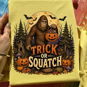 Op de afbeelding: Een lichtgeel t-shirt met een grafische print van een bruine Bigfoot die een pompoenvormige snoepemmer vasthoudt. Het ontwerp bevat een volle maan, vleermuizen, dennenbomen en pompoenen met de tekst Trick or Squatch in oranje letters.