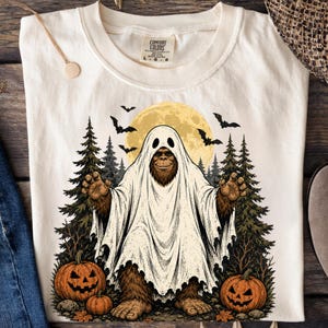May include: A cream-coloured t-shirt featuring a graphic of a brown Bigfoot creature wearing a white ghost sheet costume. The design includes a yellow full moon, black bats, evergreen trees, and two orange jack-o'-lanterns on the ground.
