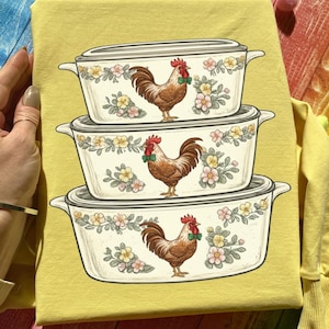 Puede incluir: Una camiseta amarilla con un diseño de tres fuentes para hornear de color crema apiladas. Cada fuente está decorada con un estampado floral y un gallo marrón con una pajarita verde. El diseño es una ilustración caprichosa.