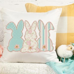 May include: White decorative pillow featuring three bunny silhouettes in pastel colors. The bunnies have polka dots, floral patterns, and stripes, with pink lace trim and flower accents. A yellow plaid pillow is in the background.