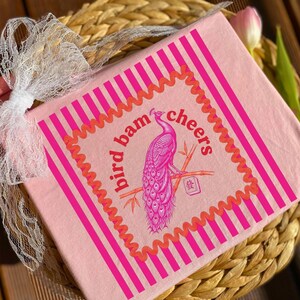 May include: A pink square item featuring a peacock design and the words "bird bam cheers". The illustration is in pink and orange, with vertical stripes and a wavy border. It is tied with a white lace ribbon and rests on a woven basket.