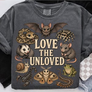 May include: A charcoal gray t-shirt featuring a graphic print of various animals including a bat, snake, spider, opossum, mouse, snail, scorpion, moth, frog, and centipede. The text LOVE THE UNLOVED is printed in large beige letters in the center.