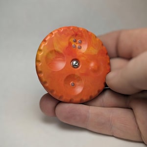 May include: A round, orange button with a marbled effect and a silver center. The button has four small holes and a textured edge. The button is held in a hand against a white background.