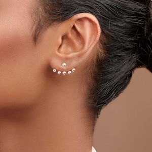 Puede incluir: Pendientes de plata con tachuelas. Una pequeña tachuela redonda está en el lóbulo de la oreja, con una fila curva de cinco tachuelas más pequeñas debajo. Los pendientes se usan en la oreja de una persona, con el pelo recogido.